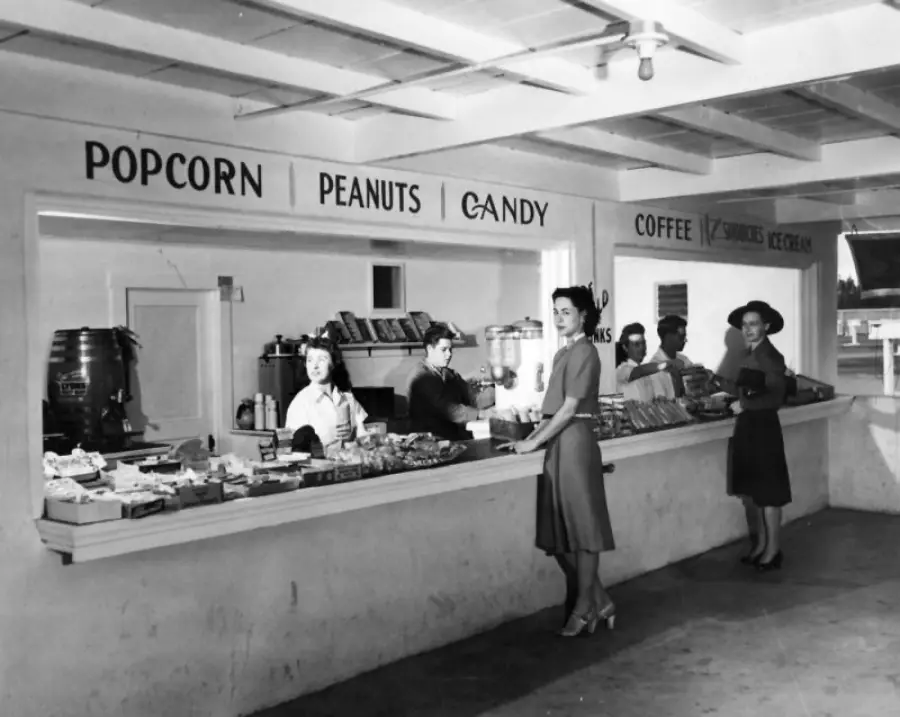 Black-and-white photo of a vintage concession stand with five women; some serve snacks like popcorn and candy, while others stand at the counter. Signs above list offerings: popcorn, peanuts, candy, coffee, sandwiches, ice cream.