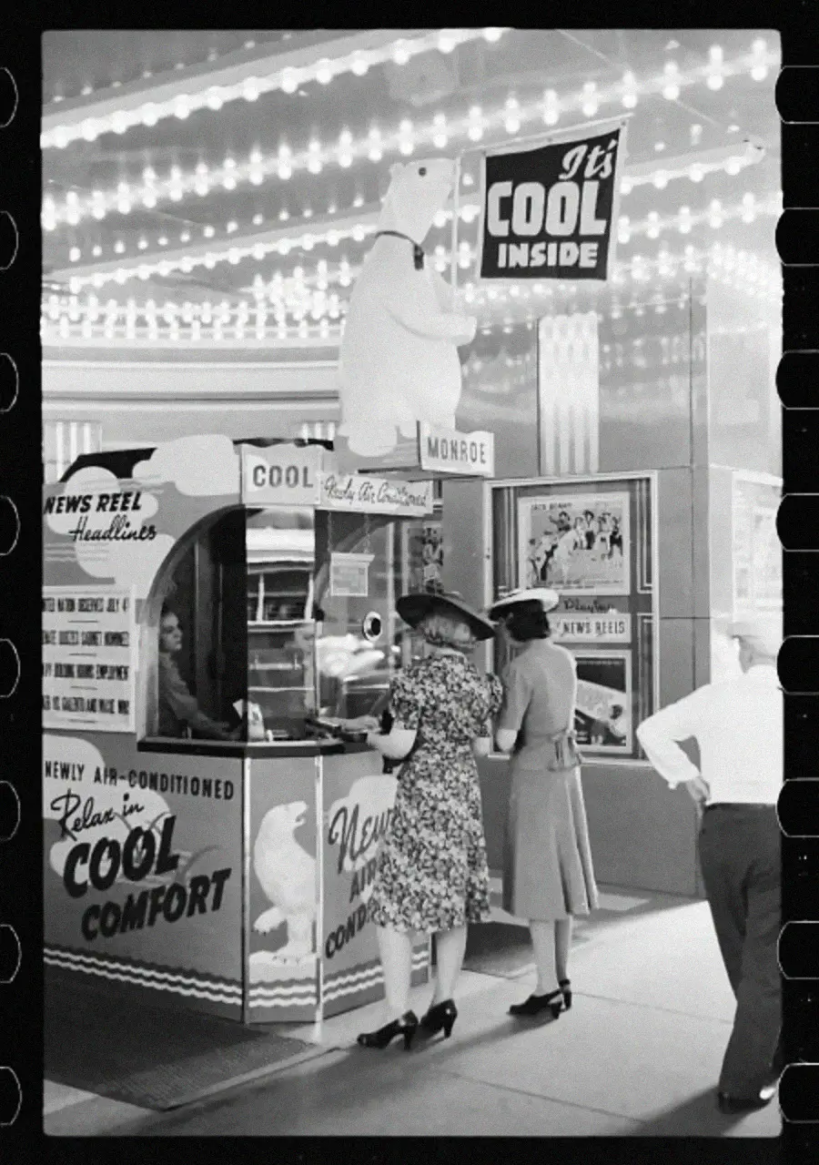 Two women stand at an outdoor movie theater ticket booth with a polar bear statue and signs reading “It’s COOL inside” and “NEWLY AIR-CONDITIONED.“ Lights decorate the entrance. A man stands nearby with hands on hips.
