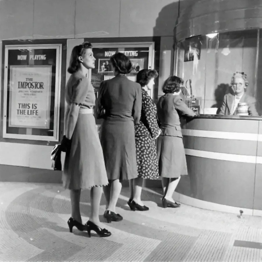 Four women stand in line at a vintage movie theater box office. A cashier serves tickets behind glass. Movie posters for “The Impostor” and “This Is The Life” are displayed on the wall.