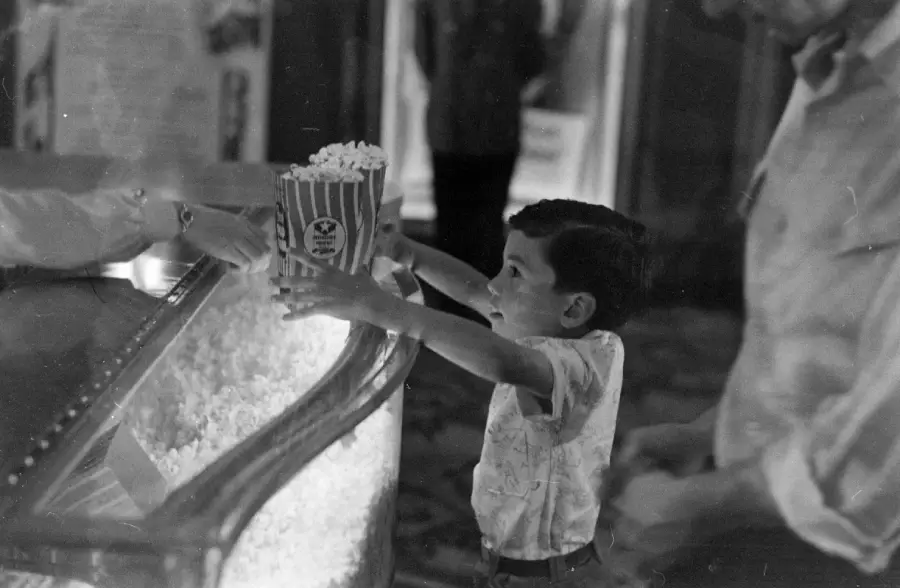 A young boy reaches up to receive a large box of popcorn at a concession stand, smiling eagerly. An adult stands beside him. The setting appears to be a theater or cinema.