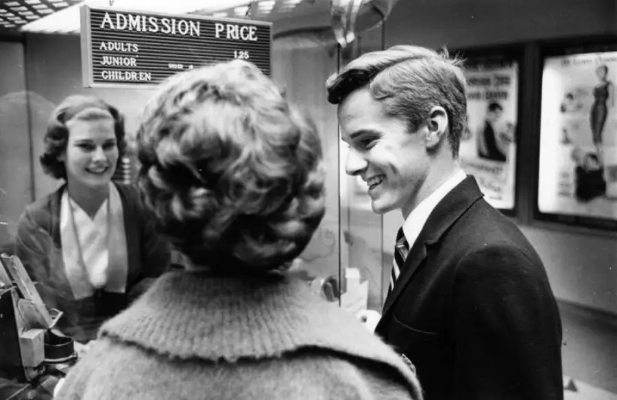 Two people stand at a movie theater ticket counter, smiling at a cashier. The admission price sign is visible above them, and movie posters are seen in the background.