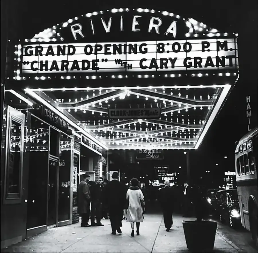 People walk toward a brightly lit theater marquee reading “Riviera Grand Opening 8:00 P.M. ‘Charade’ with Cary Grant” at night, with lights reflecting on the sidewalk and cars parked nearby.