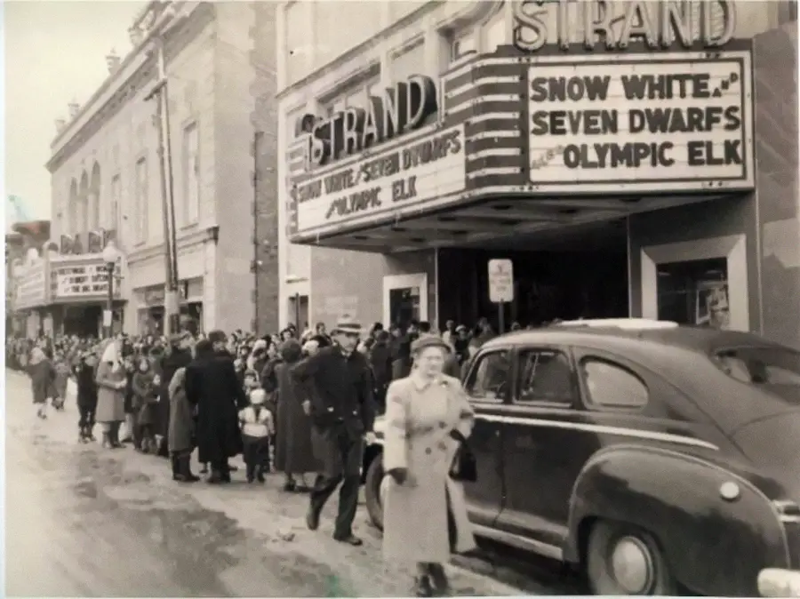 Black-and-white photo of people lined up outside a theater called “Strand”; marquee advertises “Snow White and the Seven Dwarfs” and “Olympic Elk.” Vintage cars are parked along the snowy street.