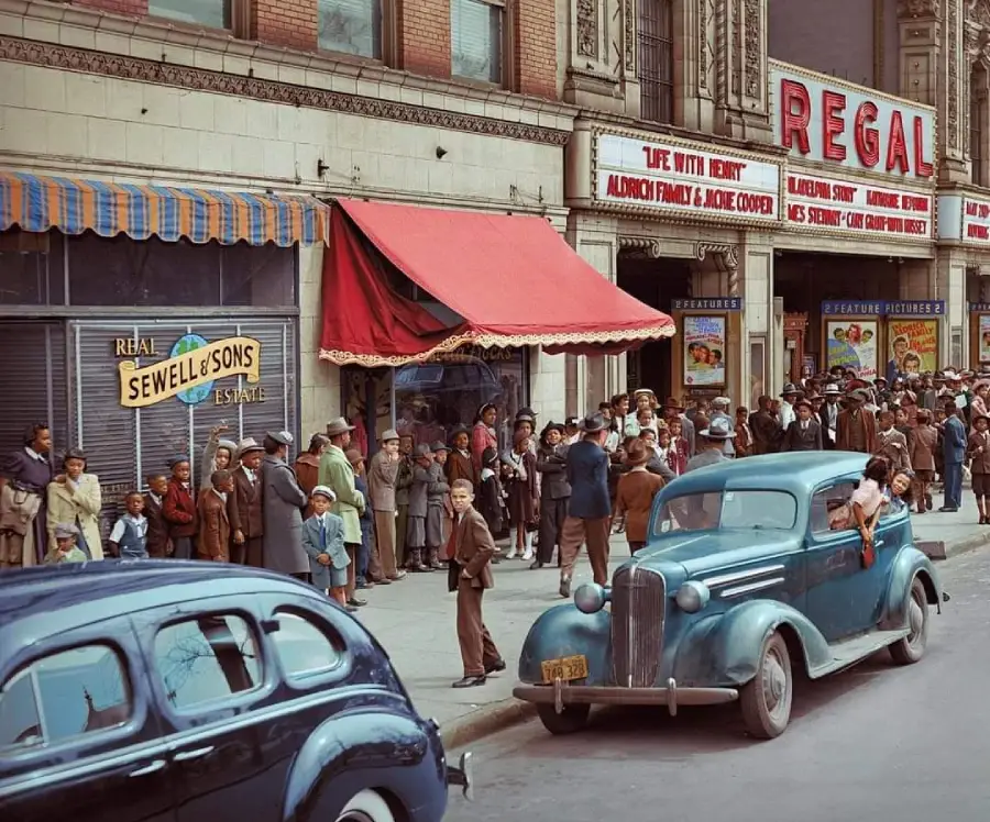 A crowd of people stands outside a theater called Regal, next to parked vintage cars. Posters advertise shows, and a sign for Sewell & Sons is visible on a neighboring storefront. The scene appears to be from the early 20th century.
