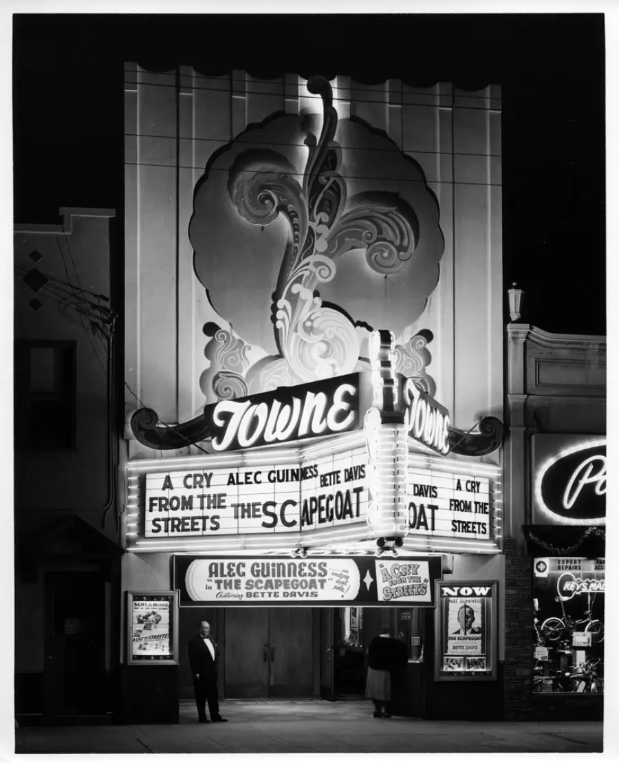 Black-and-white photo of a vintage theater at night. The marquee reads “Towne” and advertises “The Scapegoat” and “A Cry from the Streets.” Two people stand at the entrance under bright lights and ornate decor.