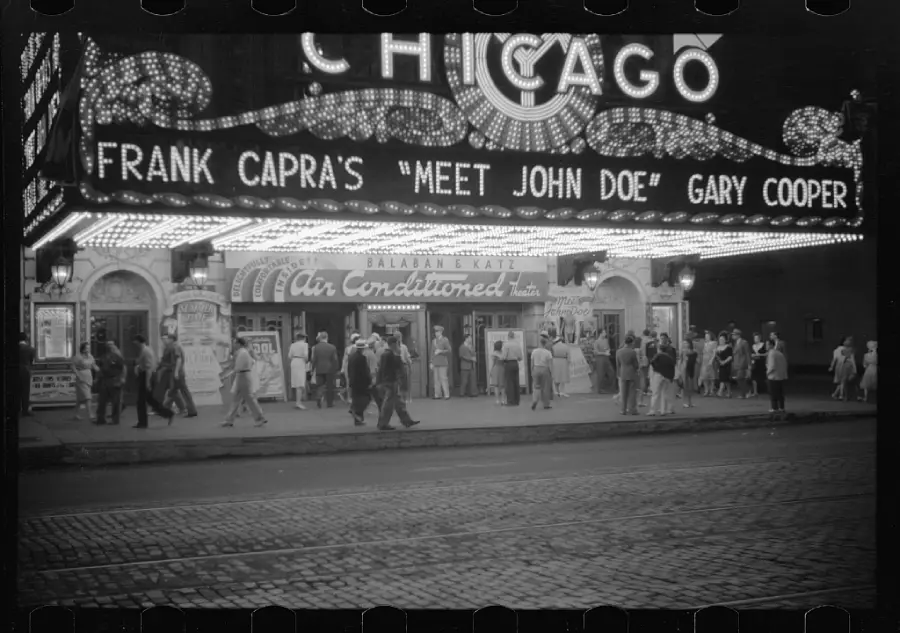 Black and white photo of a theater marquee reading “Frank Capra’s ‘Meet John Doe’ Gary Cooper” with groups of people gathered outside under bright lights at night.