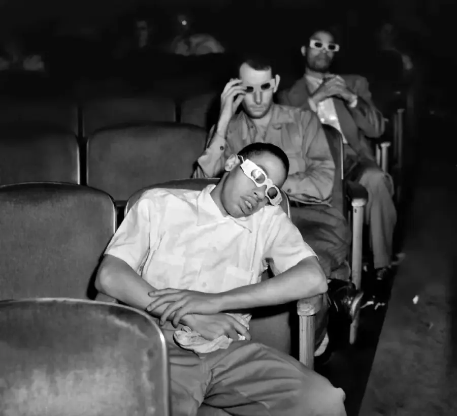 Three men sit in a nearly empty theater wearing 3D glasses; the man in front is asleep, while the two men behind him watch the screen.