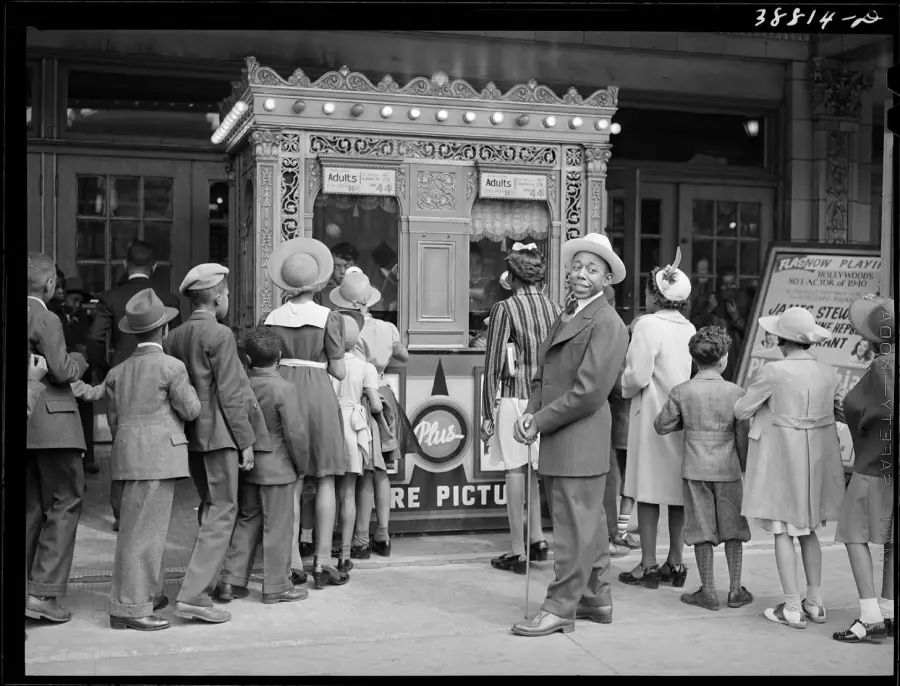 A group of people, including children and adults, waits in line at an ornate theater ticket booth. A man looks back and smiles at the camera. Everyone is dressed in vintage clothing.