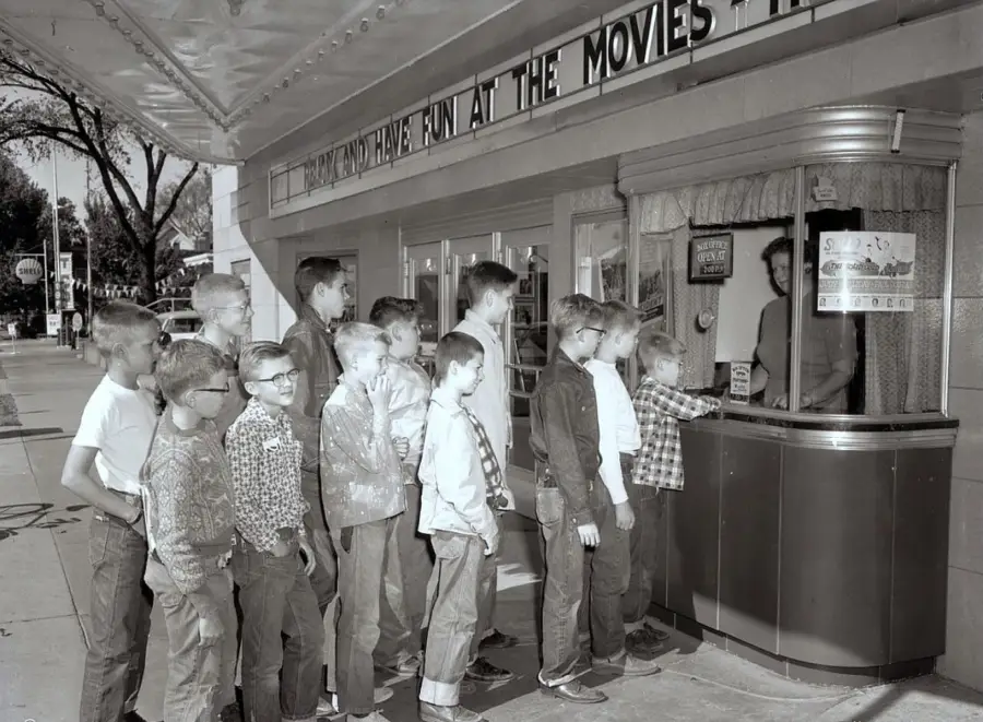 A group of boys in vintage clothing wait in line at a theater ticket booth, under a marquee that reads “HAVE FUN AT THE MOVIES.” The scene appears to be from the mid-20th century.