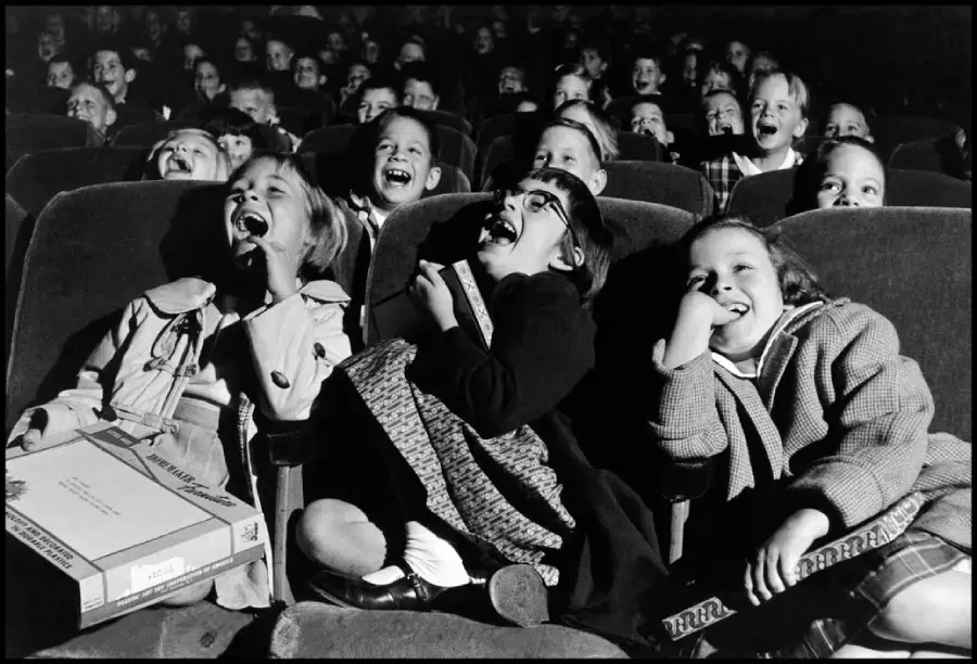 A group of children sit in theater seats, laughing joyfully at a performance or movie. Three girls in the front row are especially animated, with wide smiles and open mouths.