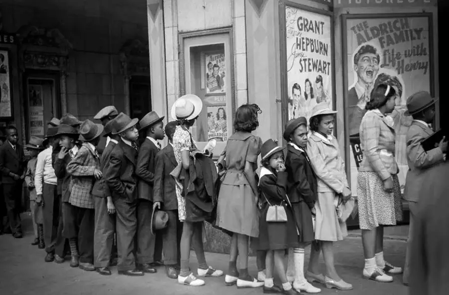 A group of children in vintage clothing stand in line outside a movie theater, waiting to buy tickets. Movie posters are visible on the wall behind them.