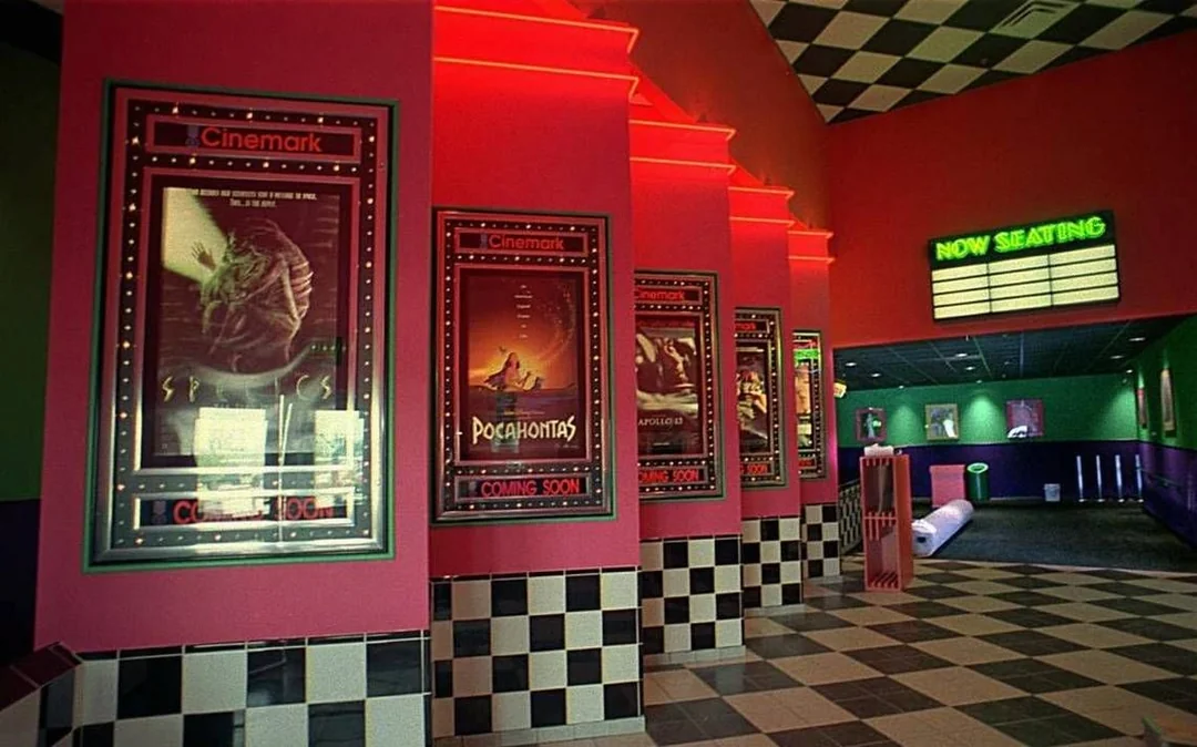 A movie theater lobby with neon red lights, checkered floors, film posters on the walls, and a “NOW SEATING” sign in the background. The décor has a retro 90s style.