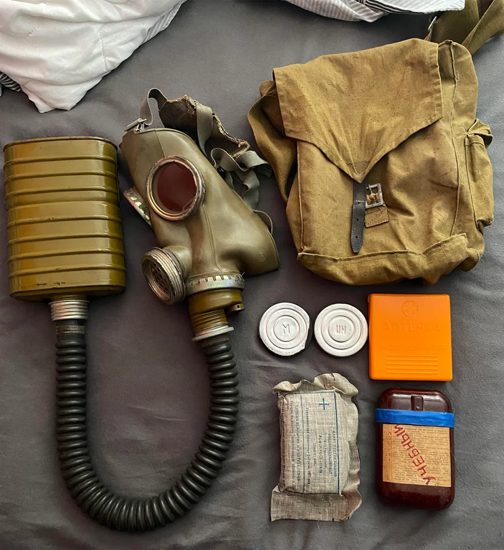 Soviet-era gas mask with filter, carrying bag, two canisters, first aid bandage, orange case, brown case, and item labeled in Russian, all laid out on a gray surface.
