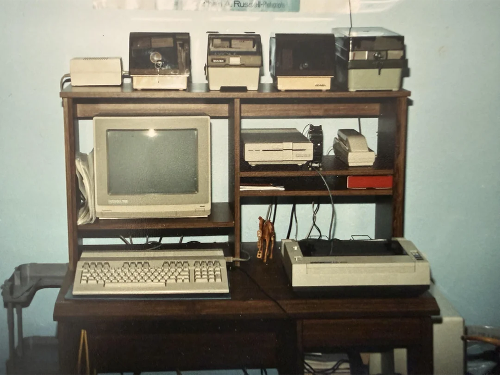 A vintage computer setup on a wooden desk with a CRT monitor, keyboard, printer, external drives, and multiple devices stacked on shelves against a light blue wall.