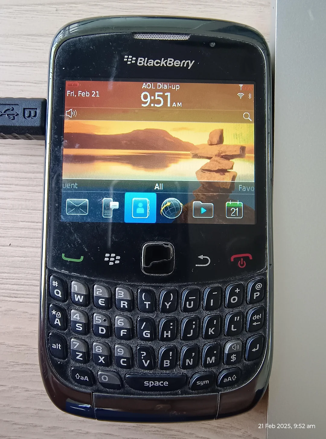 A BlackBerry smartphone with a physical QWERTY keyboard, showing the home screen at 9:51 AM on Friday, Feb 21. The screen displays icons, a sunset wallpaper, and a USB drive is plugged in on the left.
