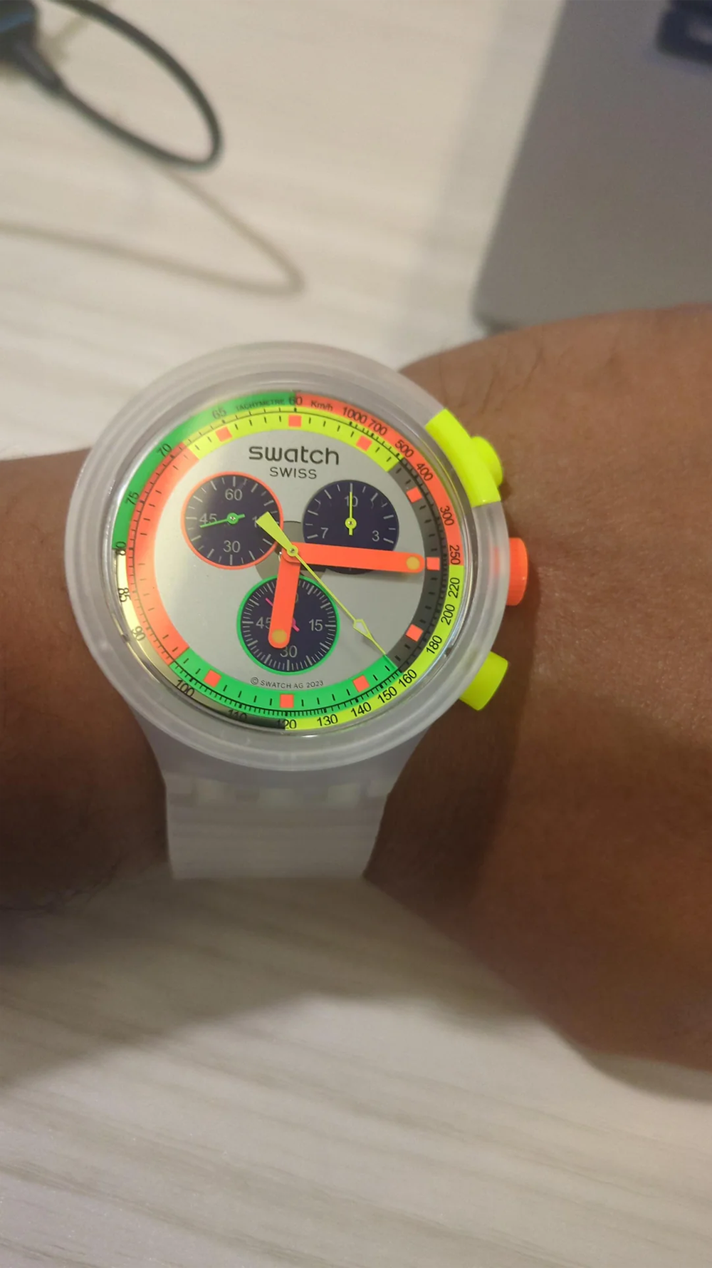 A person wearing a colorful Swatch wristwatch with a transparent band, neon green and yellow buttons, and a multicolored dial on their left wrist, on a light-colored surface.