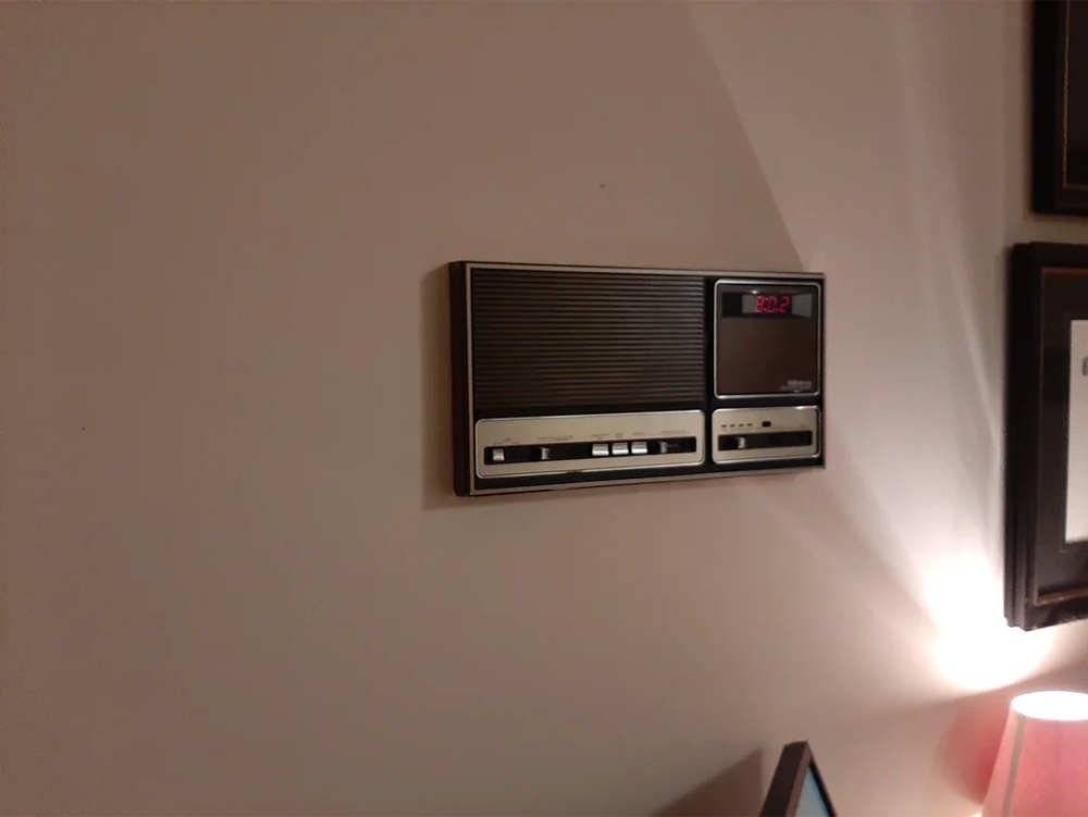A vintage wall-mounted radio and clock combo with an LED display showing 8:02, above a lit lamp and part of a framed picture on a beige wall.