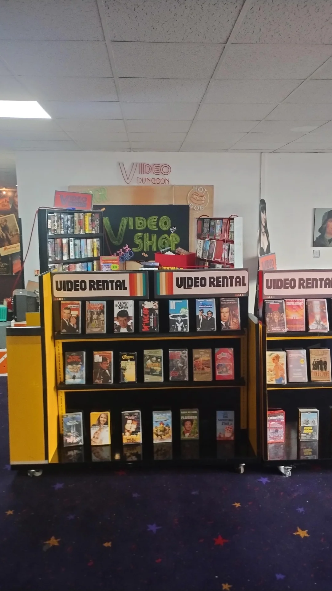 A retro video rental shop with shelves full of VHS tapes, labeled “Video Rental.” Colorful movie covers are visible. A large “Video Shop” sign and posters decorate the background walls.