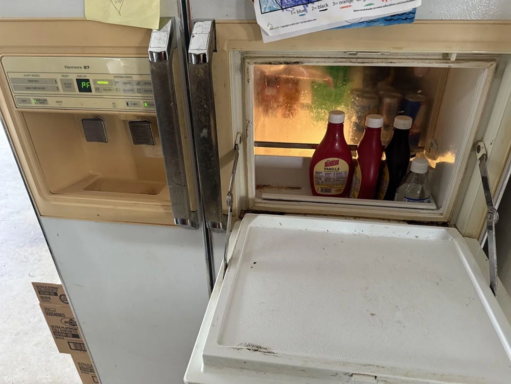 A side-by-side refrigerator with its ice dispenser door open, revealing bottles of condiments and drinks stored inside the ice compartment. The fridge is off-white and appears to be older in style.
