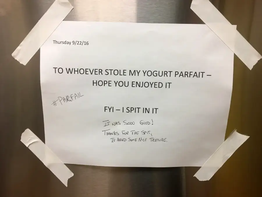 A taped sign reads: “To whoever stole my yogurt parfait—hope you enjoyed it. FYI—I spit in it,” with handwritten notes underneath and hashtags, all on a metallic surface.