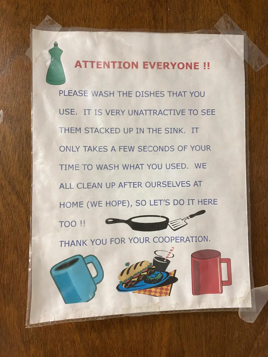 A printed sign taped to a wooden surface asks people to wash their dishes, with colorful images of mugs, a plate with food, utensils, and a green dish soap bottle. The sign has text in blue and red.