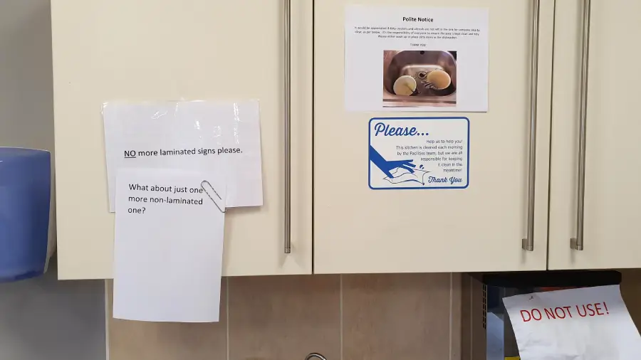Several signs are attached to cabinet doors. One sign requests no more laminated signs, while another below humorously asks, “What about just one more non-laminated one?” Other signs include kitchen notices and a “DO NOT USE!” label.