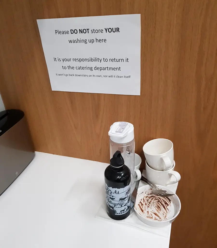 A sign on a wood panel reads “Please DO NOT store YOUR washing up here.” Below it, dirty cups, a plate, and water bottles are stored there anyway.