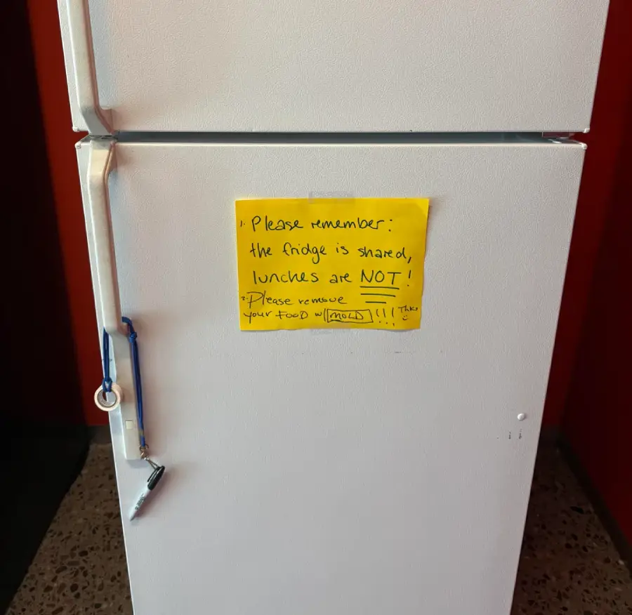 A white fridge with a lock on the handle and a yellow note reminding people the fridge is shared but lunches are not; it asks to remove old food.