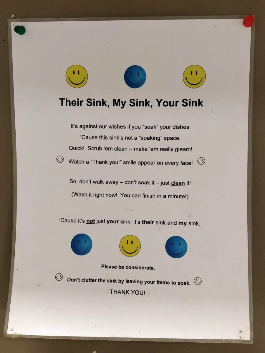 A printed sign with yellow and blue smiley faces reminds people not to leave dishes soaking in the sink and to clean them right away. It emphasizes consideration for others and keeping the sink tidy.