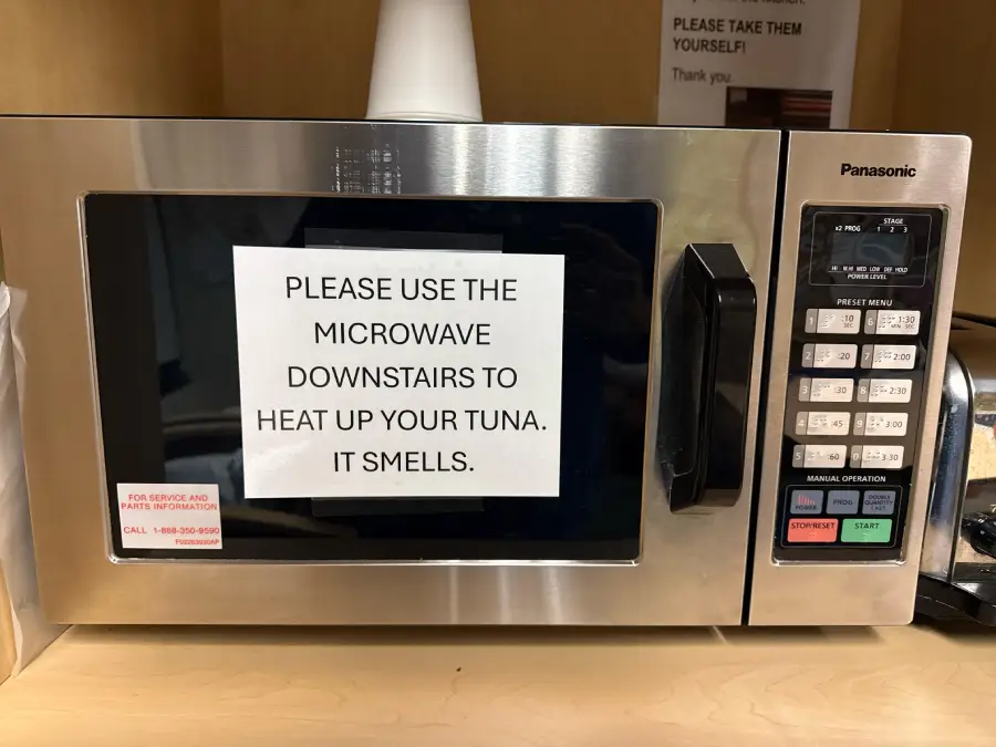 A microwave with a sign reading, “PLEASE USE THE MICROWAVE DOWNSTAIRS TO HEAT UP YOUR TUNA. IT SMELLS.” displayed on the door.