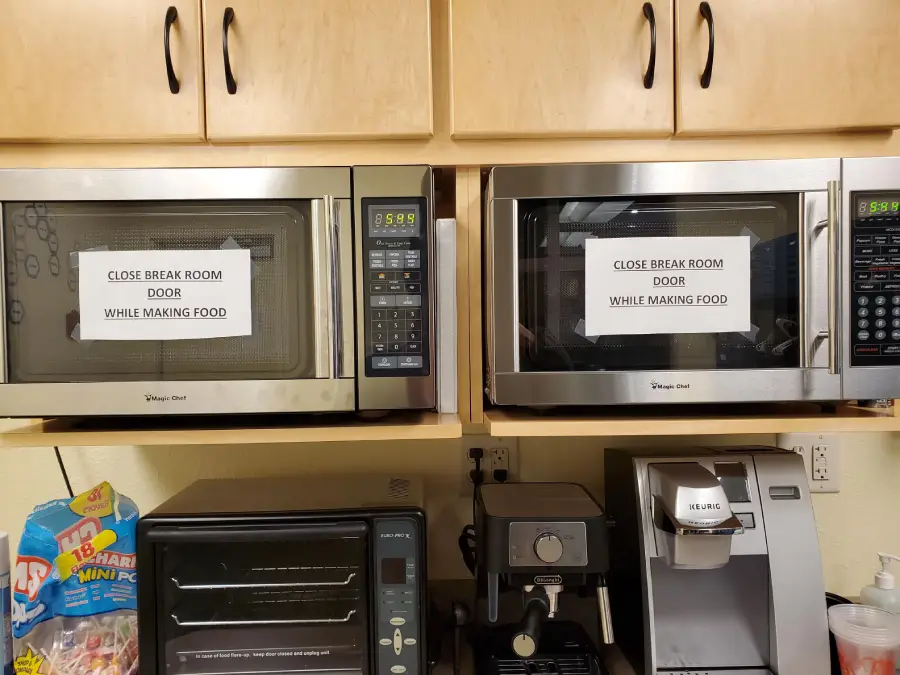 Two microwaves with identical signs reading “CLOSE BREAK ROOM DOOR WHILE MAKING FOOD,” above a coffee maker, chips, and snacks in an office kitchen setting.