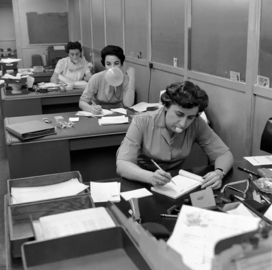 Three women work at desks in an office. The woman in front is writing, another blows bubble gum, and the third works in the background. Papers and office supplies are on the desks.