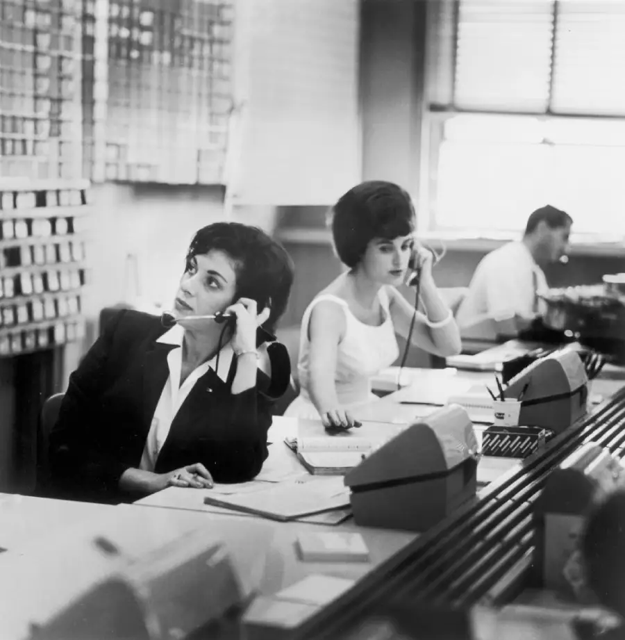 Two women wearing headsets sit at desks with papers and early office equipment, working in a vintage office. A man is seated in the background. The setting appears to be a busy mid-20th-century workplace.