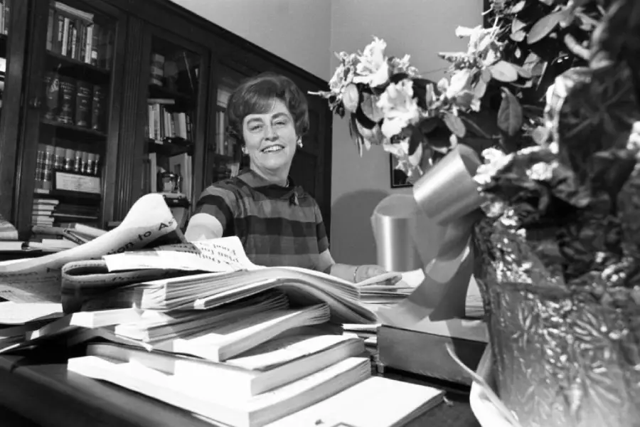 A woman sits at a desk covered with papers and books, smiling at the camera. There are bookshelves behind her and a vase with flowers and a ribbon in the foreground.