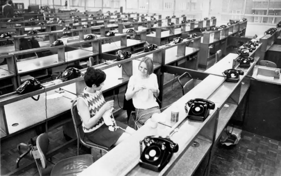 Two women sit and knit at a long desk lined with black rotary phones in a large, mostly empty call center or office space. Rows of empty desks and chairs fill the background.