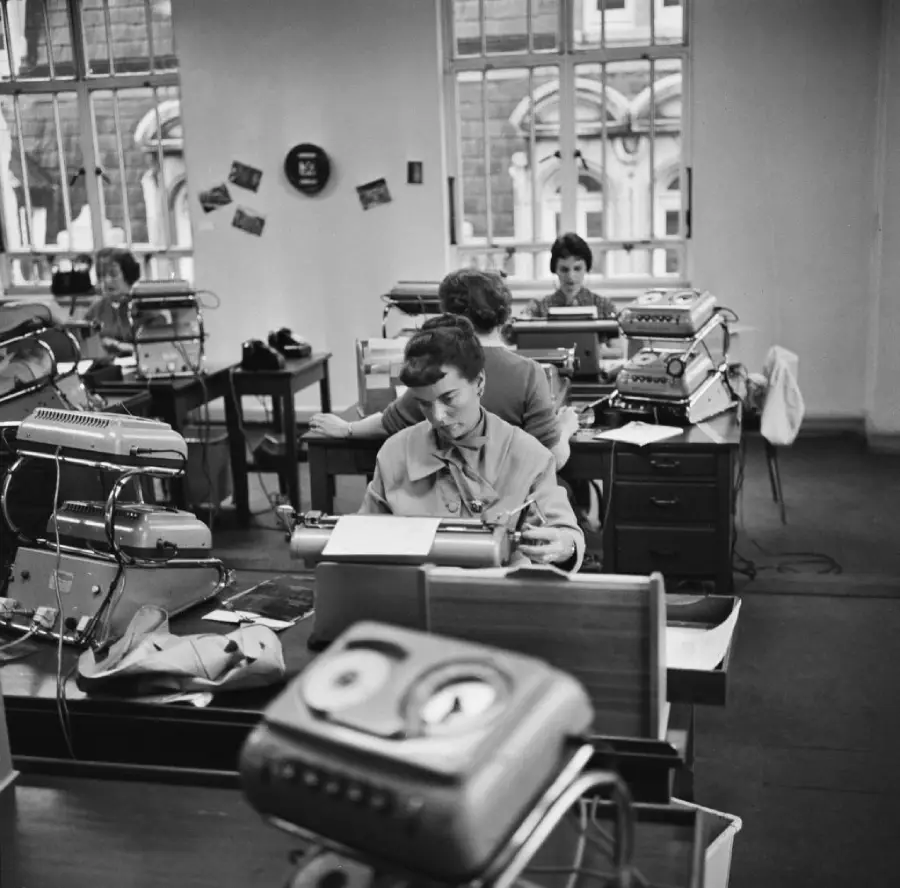 Black-and-white photo of women working at desks with typewriters and paperwork in an office with large windows, likely from the mid-20th century. The room looks busy and organized.
