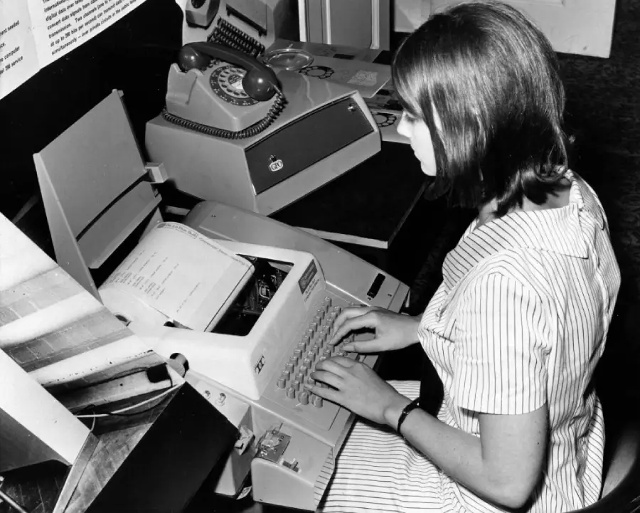 A woman in a striped dress types on an old-fashioned typewriter, with a rotary phone and papers nearby, in a black and white photo.