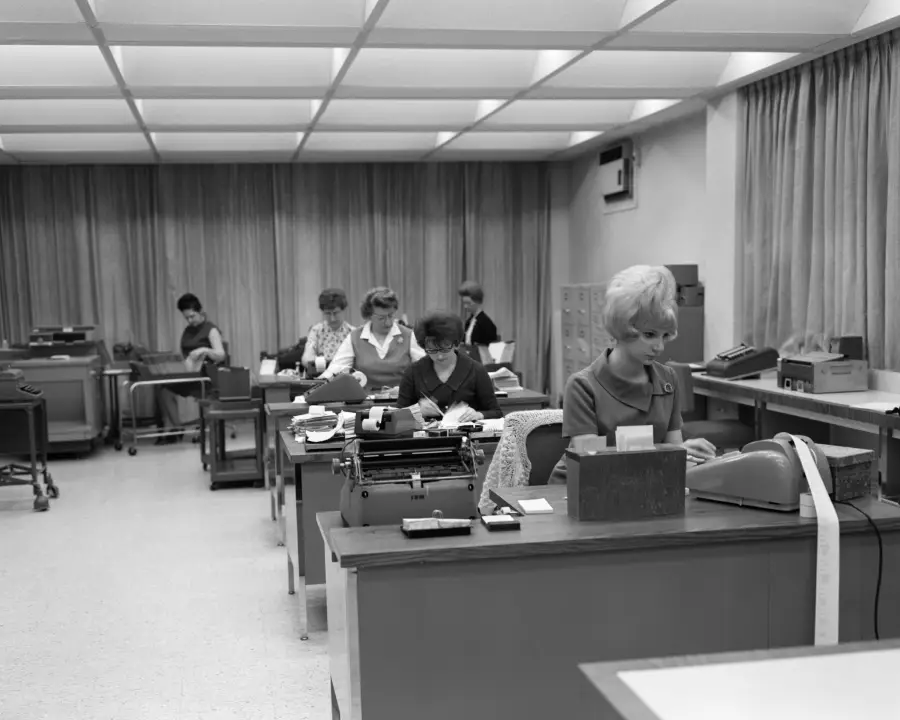 Black and white photo of women working at desks with typewriters and calculators in an office, likely from the 1960s. The room has large windows, curtains, and ceiling lights.