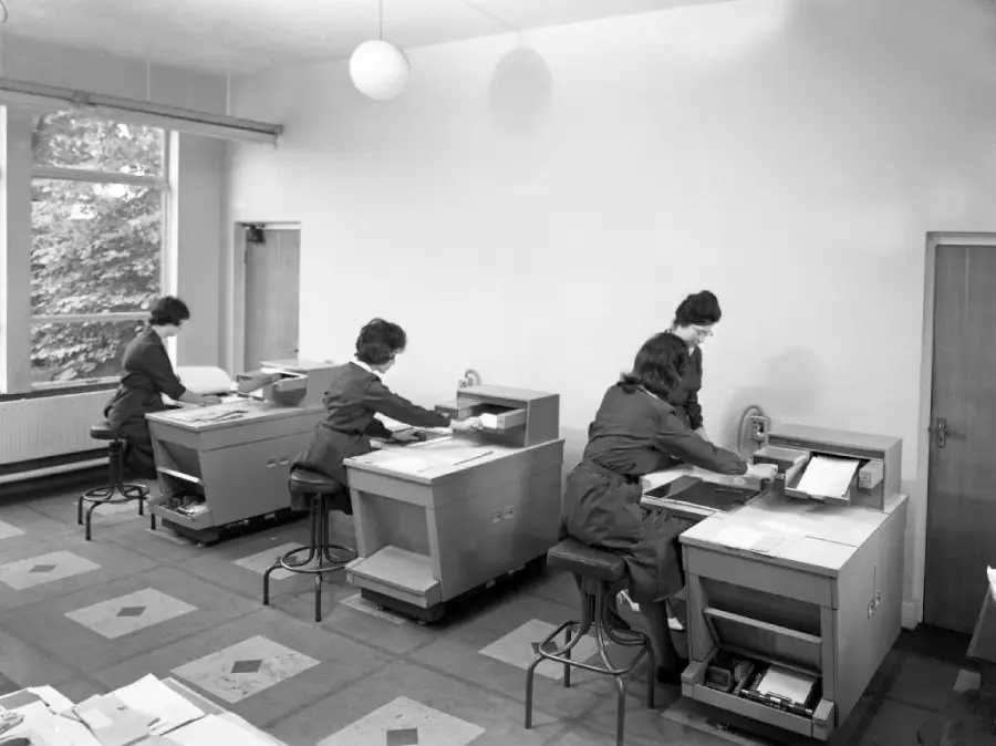 Four women work at separate desks in a bright, tidy office with large windows and scattered papers. Each is focused on paperwork, dressed in similar outfits, with minimal decor and a modern, mid-20th-century style.