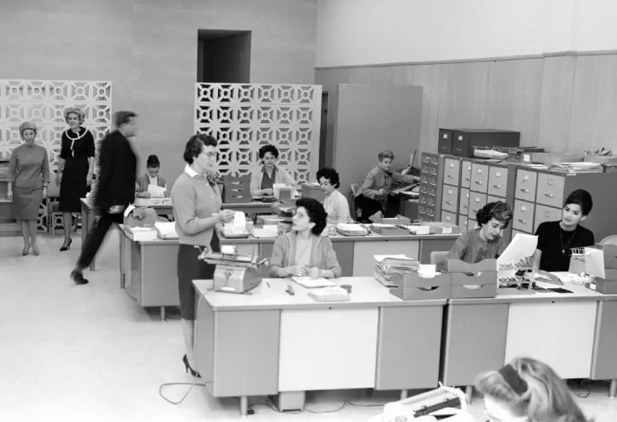 Black-and-white photo of a busy mid-century office with women working at desks, typing, filing, and talking. A man walks past in the background. Several filing cabinets and decorative dividers are visible.