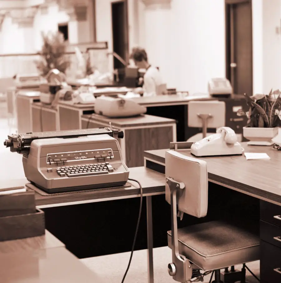A sepia-toned photo of a vintage office with desks, typewriters, rotary phones, and papers. A person sits at a desk in the background, and there are plants on some desks.