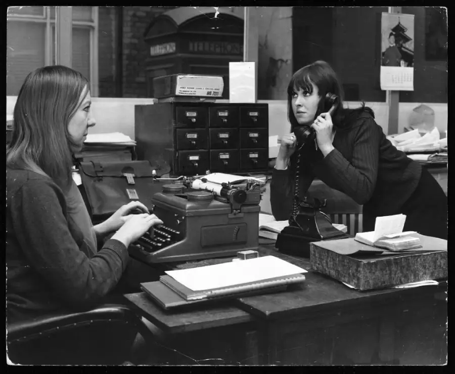 Two women work at an office desk; one types on a typewriter while the other talks on a rotary phone, leaning forward. The desk is cluttered with papers and office supplies.