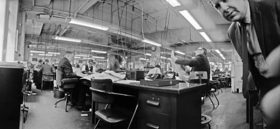 Black and white image of a busy office with people working at desks, papers scattered, telephones, and fluorescent lighting overhead. A man in a suit is close to the camera on the right side.