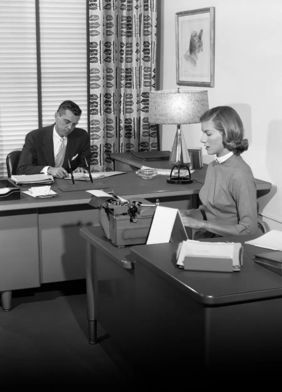 A man in a suit writes at his desk behind a woman using a typewriter at another desk. The office has mid-century furniture, patterned curtains, a table lamp, and framed art on the wall.