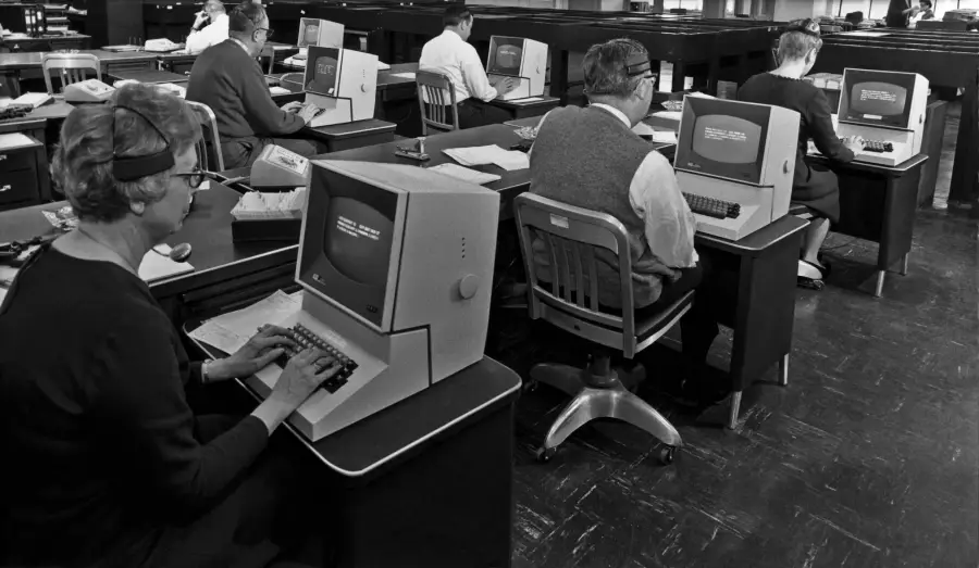 Black-and-white photo of people sitting at desks typing on vintage computer terminals in an office, likely from the mid-20th century. Each person faces a bulky monitor with white text on the screens.