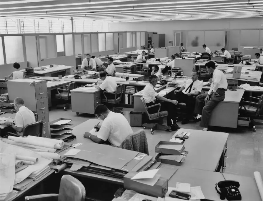 Black-and-white photo of a large, busy 1960s office with men in white shirts working at desks, talking, and reviewing papers in an open-plan space with overhead lights.