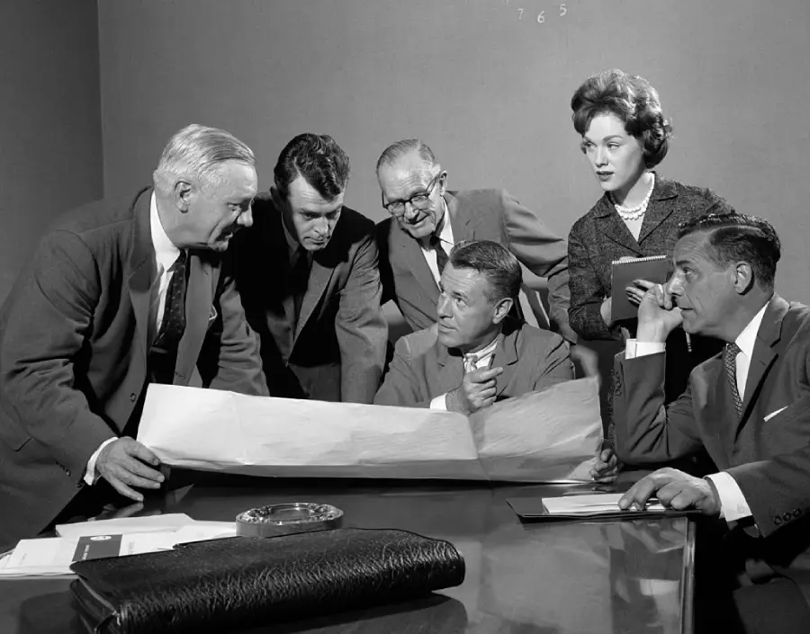 Six professionals in business attire gather around a table, intently examining a large blueprint or document; one woman stands holding a notepad while the others sit or lean forward, engaged in discussion.