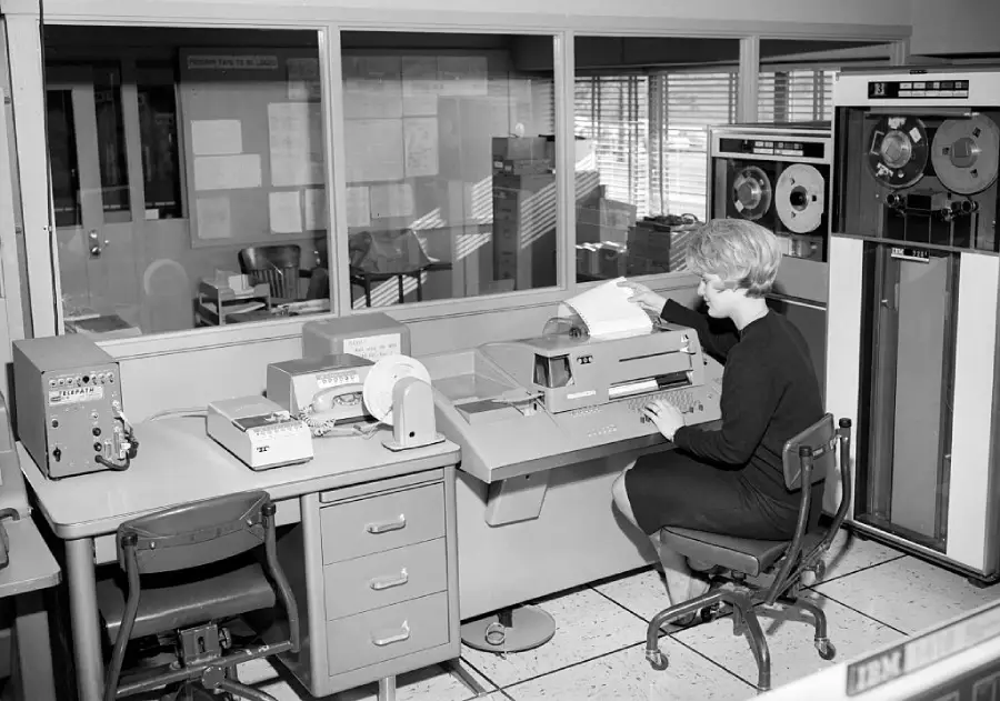 A woman in a dress operates a vintage computer workstation with paper printouts and large tape drives in a mid-20th century office setting.