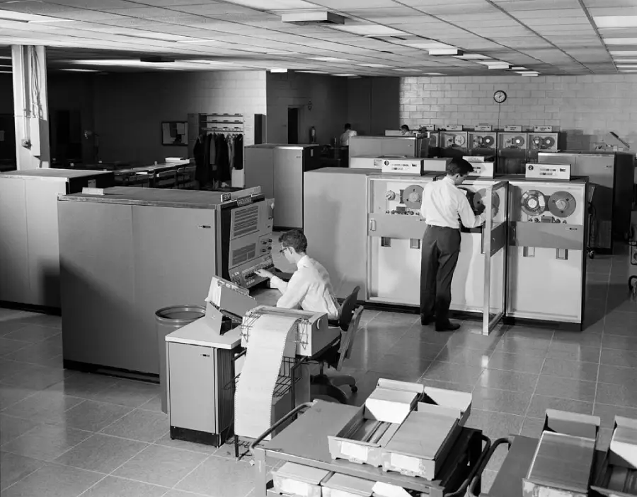 Two people operate large vintage computers in a spacious room, with one seated at a console and another using reel-to-reel tape machines. Boxes of punch cards sit nearby. The scene is from an earlier era of computing technology.
