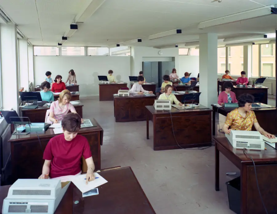 A group of women work at desks with typewriters and papers in a bright, spacious office with large windows and simple decor, suggesting a scene from the 1970s.